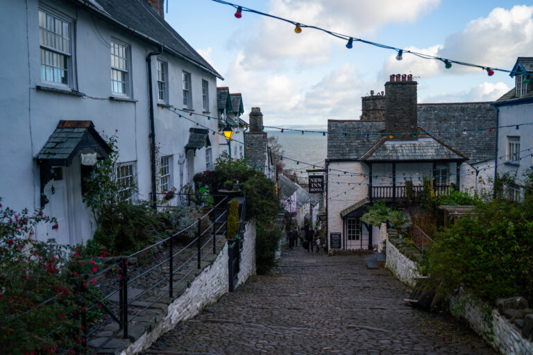 Steep cobbled street in Clovelly village lined with white cottages, string lights overhead, and a view toward the sea in the distance.