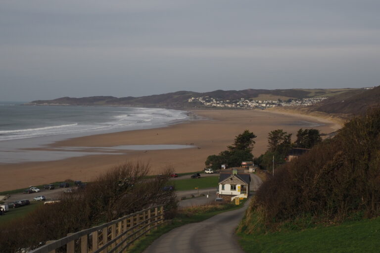 Panoramic view of Woolacombe Beach in Devon, showing a wide sandy shoreline, gentle ocean waves, rolling hills, and a seaside village in the distance under soft light.