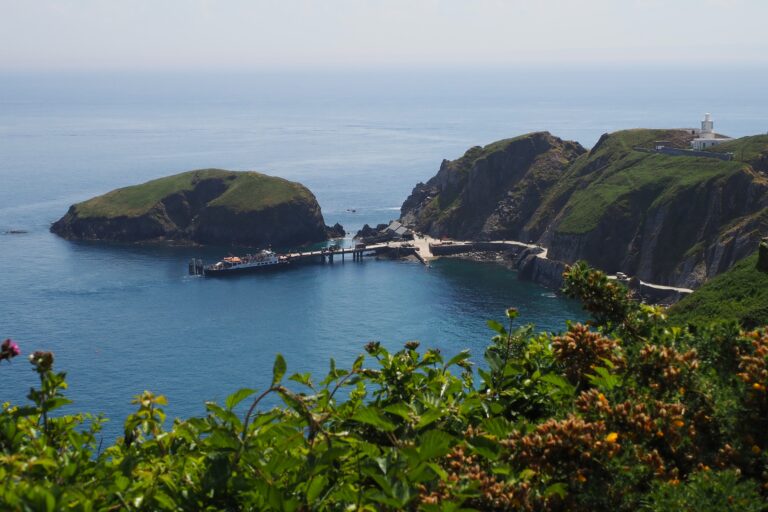 MS Oldenburg ferry docked at Lundy Island pier, surrounded by rugged cliffs and calm blue sea, with a lighthouse visible on the headland.
