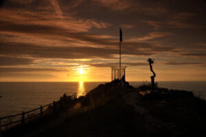 Silhouette of Kate Frolov memorial statue and flagpole against orange sunset sky.
