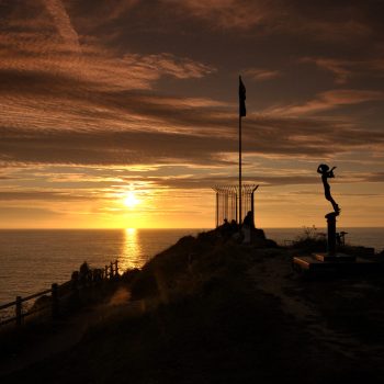 Silhouette of Kate Frolov memorial statue and flagpole against orange sunset sky.